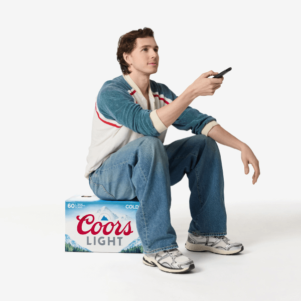 Man sitting on a case of Coors Light beer, holding a remote control, wearing jeans and sneakers against a white background.