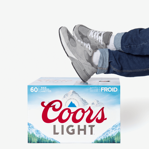 Personne portant un jean et des baskets, les pieds reposant sur un pack de bière Coors Light, sur fond blanc.