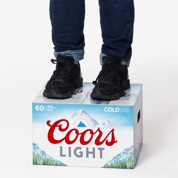 Person wearing jeans and black shoes standing on top of a Coors Light beer case against a white background.