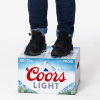 Personne portant un jean et des chaussures noires, debout sur un pack de bière Coors Light, sur fond blanc.